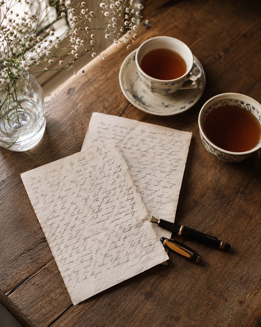 Two handwritten letters, a black and gold fountain pen, and two cups of tea sit on a wooden table next to a glass vase with white flowers.
