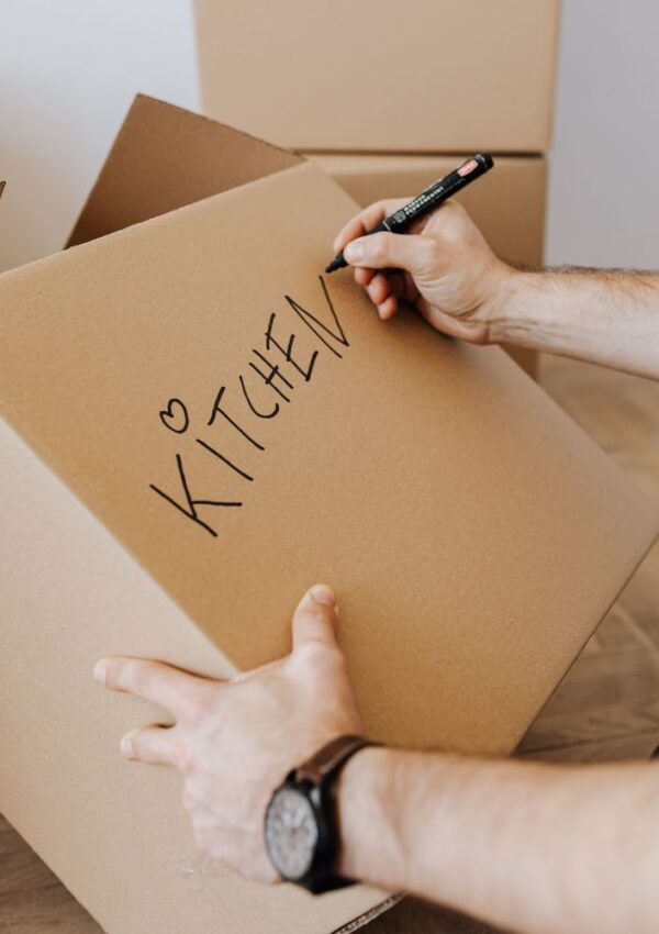 A person writes KITCHEN with a heart on a cardboard box using a black marker, preparing for a move. Other packed boxes are visible in the background.
