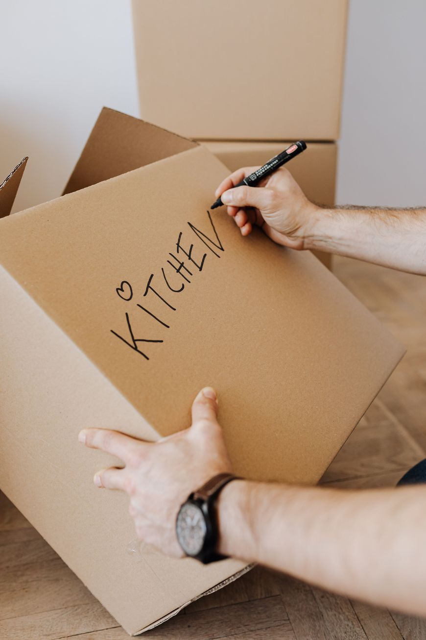 A person writes KITCHEN with a heart on a cardboard moving box using a black marker. Stacked boxes and a wooden floor are visible in the background.