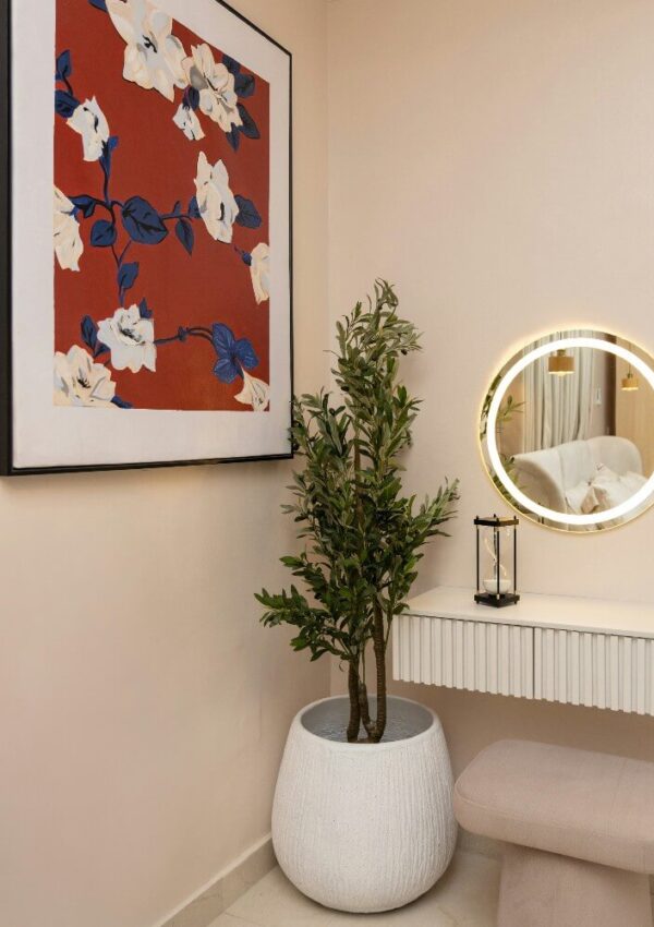 A modern vanity corner with a round lighted mirror, small bench, potted plant, and a large floral artwork on a beige wall.