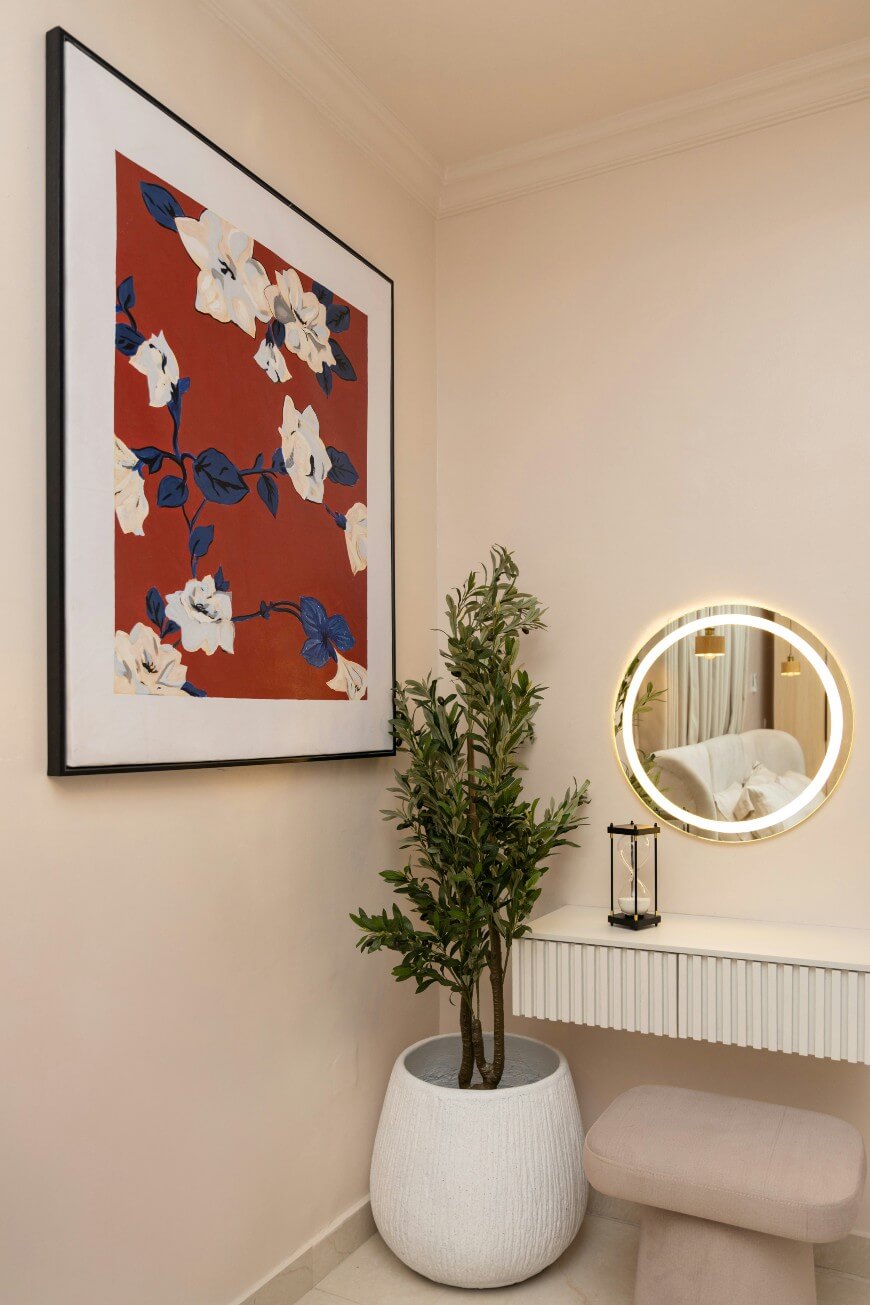 A small vanity area features a round mirror with a glowing light, a potted plant on the floor, a beige stool, and a large floral wall art with white flowers and dark leaves on a red background.