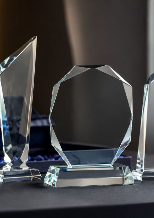 Three clear glass award trophies with geometric shapes sit on a dark table, positioned in front of a soft-focus background. The trophies reflect light and have distinct angular cuts and polished surfaces.