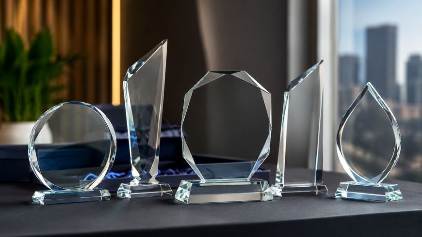 Five clear glass trophies of various geometric shapes are displayed on a dark table, with an office window and city buildings visible in the blurred background.