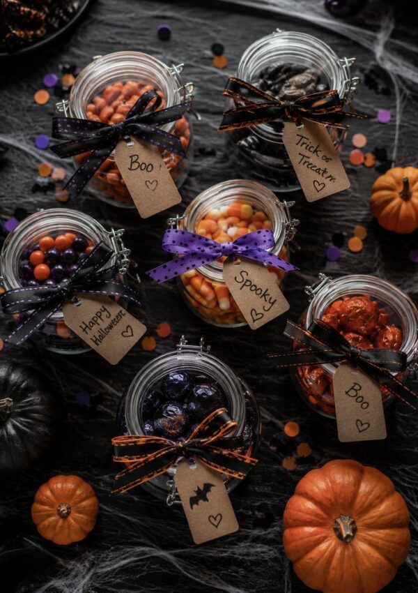 Five glass jars filled with Halloween candy, decorated with ribbons and tags saying Boo, Spooky, Trick or Treat, and Happy Halloween. Surrounded by mini pumpkins and confetti on a dark surface.