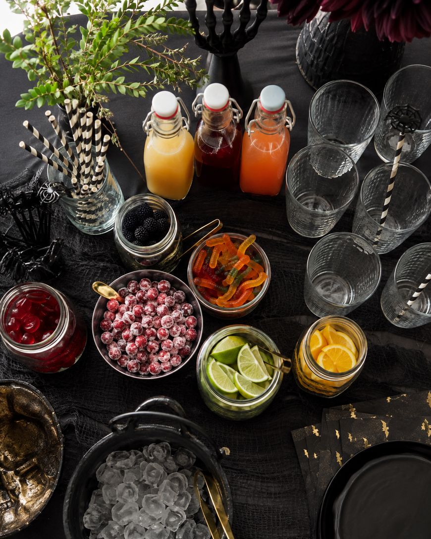 Overhead view of a drink station with glass bottles of juice, jars of fruit, candy, ice, glasses, decorative straws, and gold-rimmed napkins on a black tablecloth, accented with flowers and greenery.