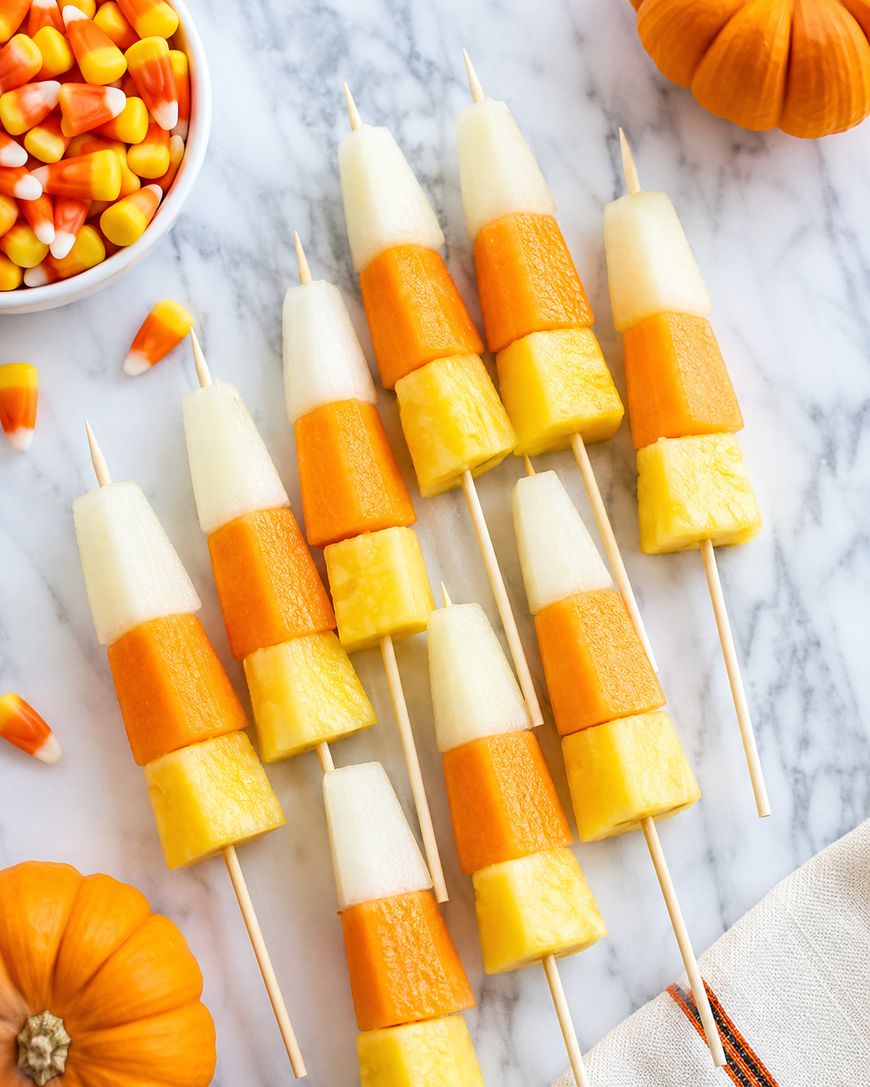 Fruit skewers on a marble surface, each with pineapple, cantaloupe, and pear pieces arranged to mimic candy corn. A bowl of candy corn, a mini pumpkin, and a napkin are also visible.