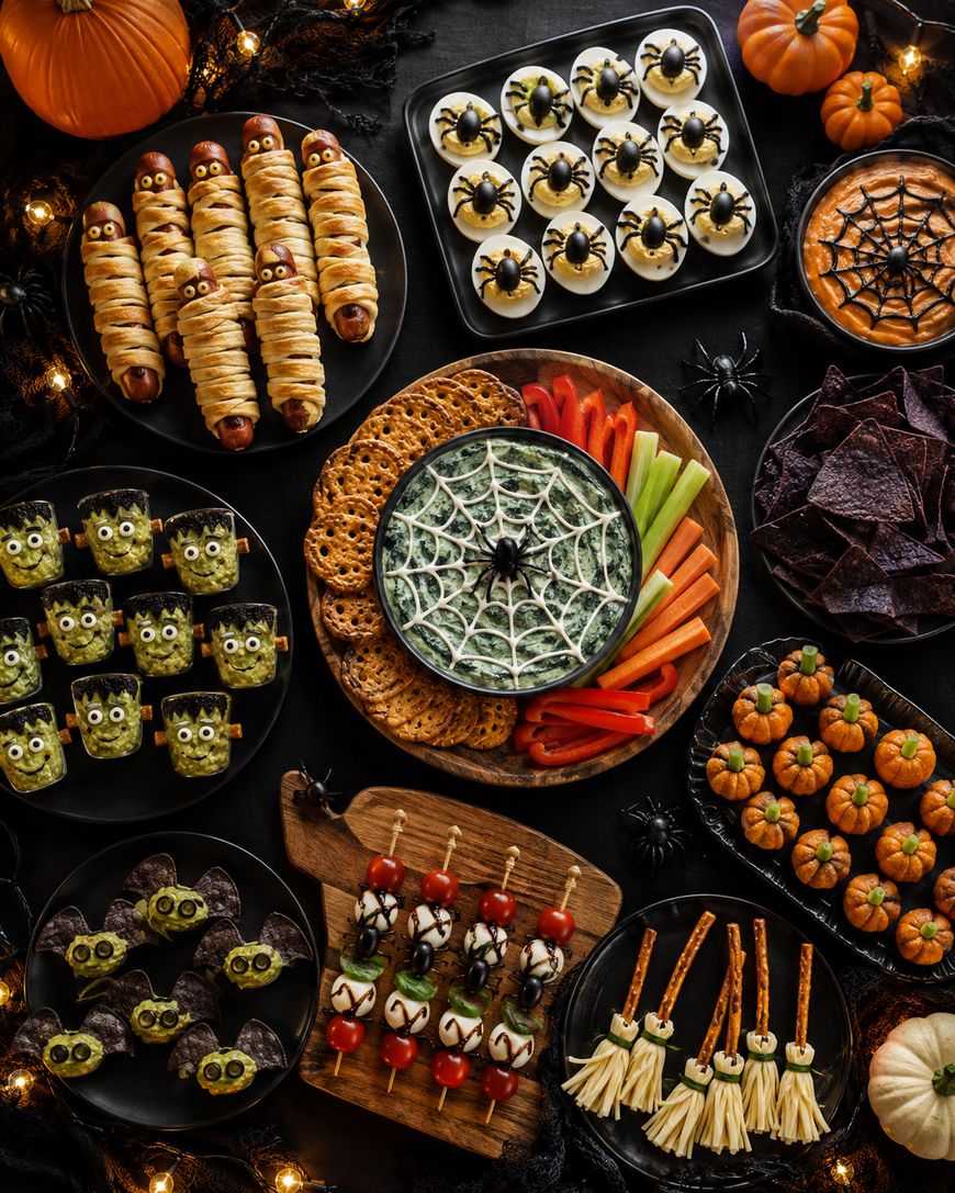 A Halloween-themed snack spread with mummy hot dogs, deviled eggs decorated as spiders, veggie platter with spiderweb dip, monster rice treats, mini pumpkin desserts, witch broomsticks, and other spooky treats.