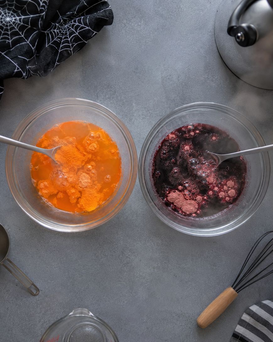 Two bowls with orange and dark purple bubbling liquids being mixed with spoons sit on a gray surface, surrounded by kitchen utensils, a tea kettle, and a black cloth with a white spiderweb pattern.