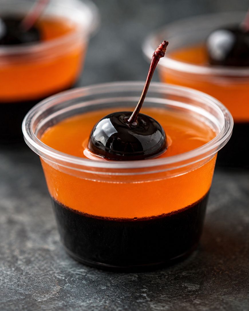 A clear plastic cup holds a two-layered dessert with an orange top, dark bottom, and a glossy cherry with stem on top. Two similar cups are blurred in the background.