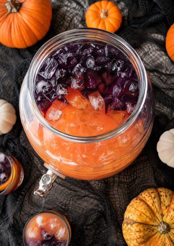 A glass drink dispenser filled with orange and purple iced punch is surrounded by small pumpkins and gourds on a black cloth, with two glasses of the punch nearby.