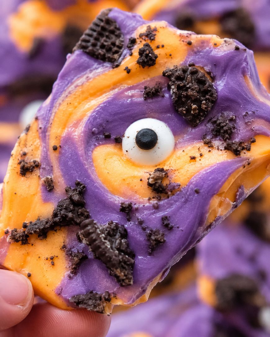 A hand holds a piece of colorful Halloween bark with swirled orange and purple chocolate, a candy eye, and crumbled cookies on top.