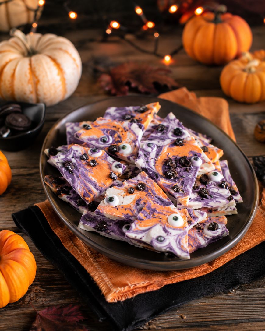 A plate of Halloween bark with purple, orange, and white swirls, candy eyes, and cookie bits sits on napkins. Mini pumpkins, lights, and autumn leaves decorate the wooden table.