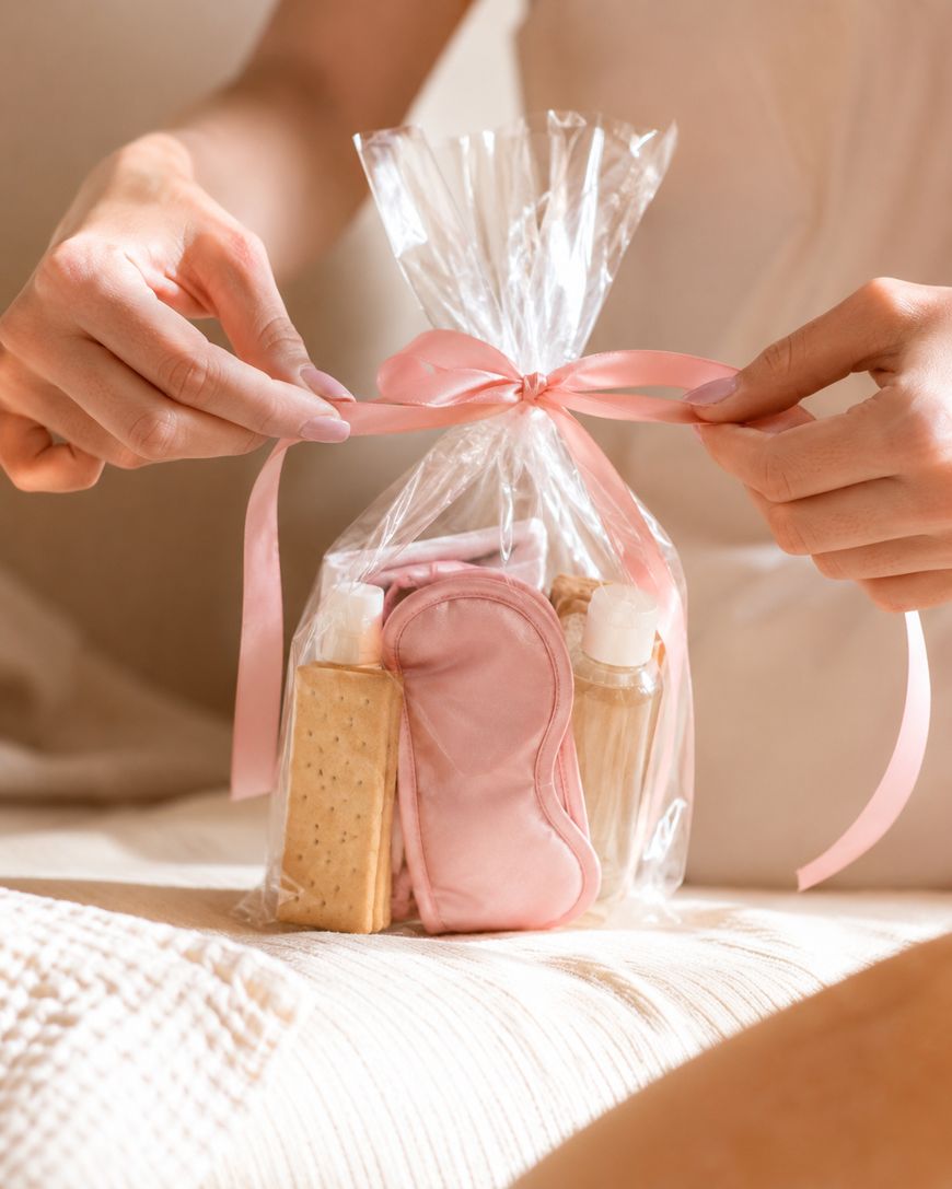 A person ties a pink ribbon around a clear gift bag containing an eye mask, snacks, and small bottles, sitting on a bed.