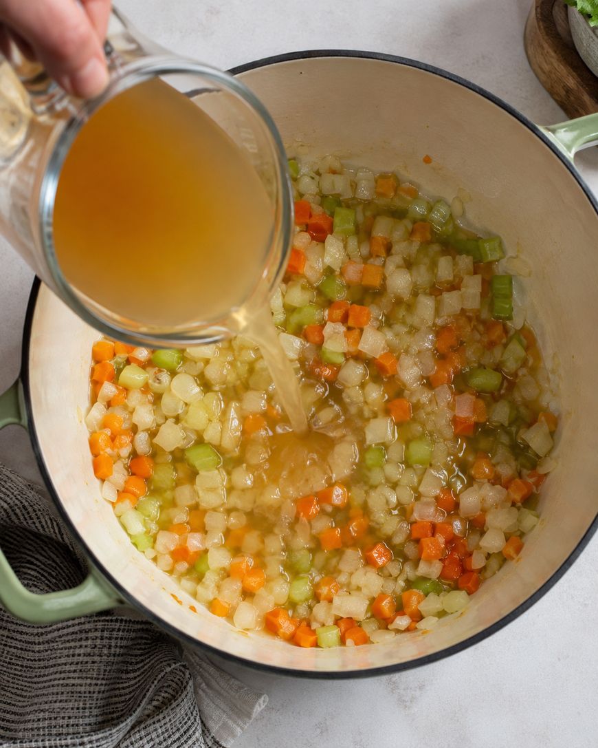 Broth being poured into a pot of sauteed onion, celery, and carrot.