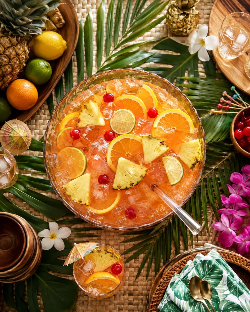 A tropical punch bowl with ice, orange slices, pineapple wedges, cherries, and lime slices, surrounded by fruits, glasses, flowers, and palm leaves on a woven mat.