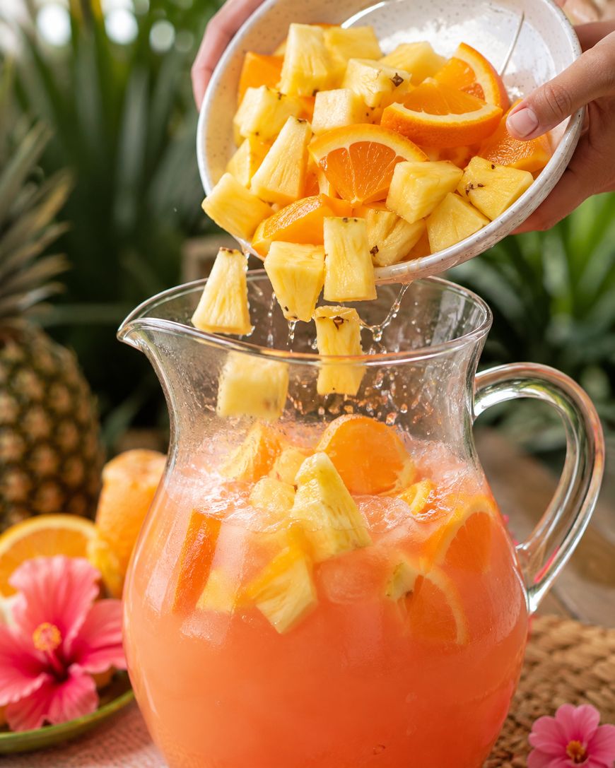 A hand pours fresh pineapple and orange pieces into a glass pitcher filled with a pink tropical drink. Pineapples, oranges, and a hibiscus flower are in the background.