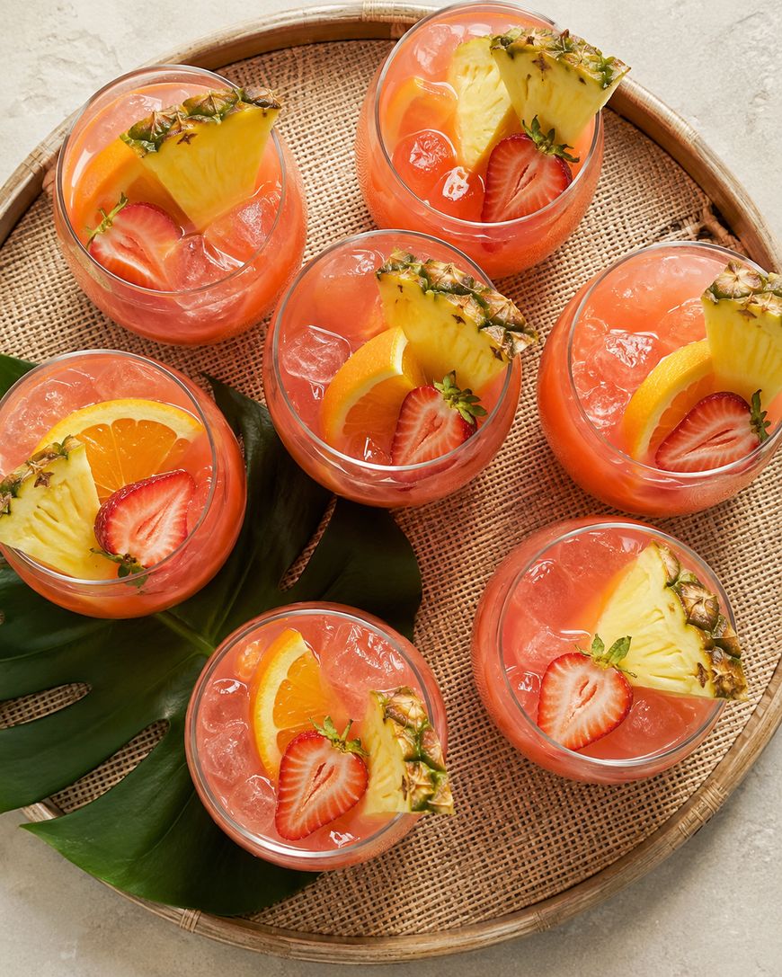 Seven glasses of pink tropical punch with ice, garnished with pineapple slices, orange wedges, and strawberries, arranged on a woven tray with a green leaf underneath.