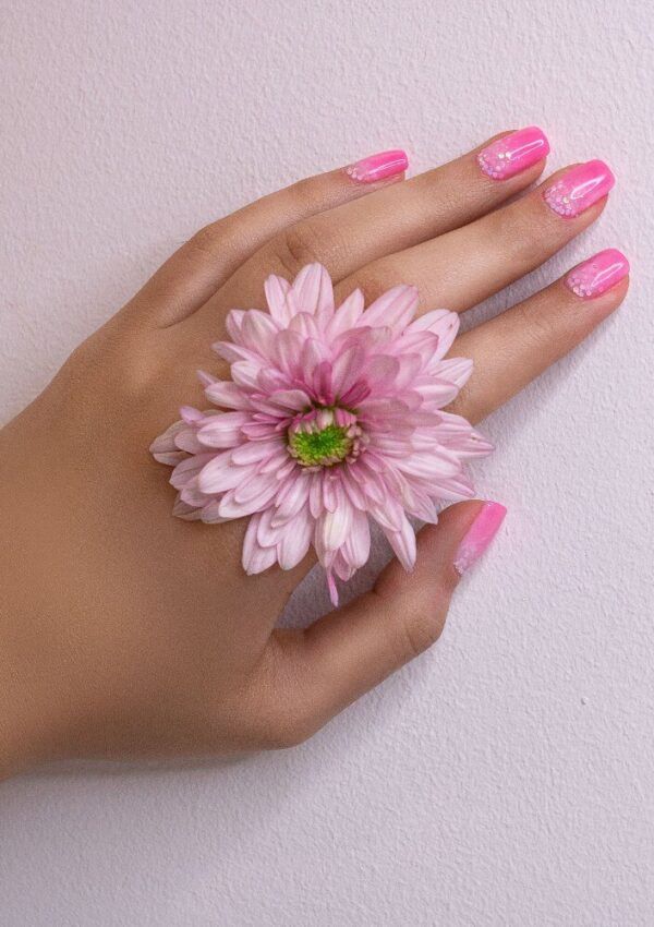 A hand with pink manicured nails rests on a light surface, with a pink flower placed between the index and middle fingers.