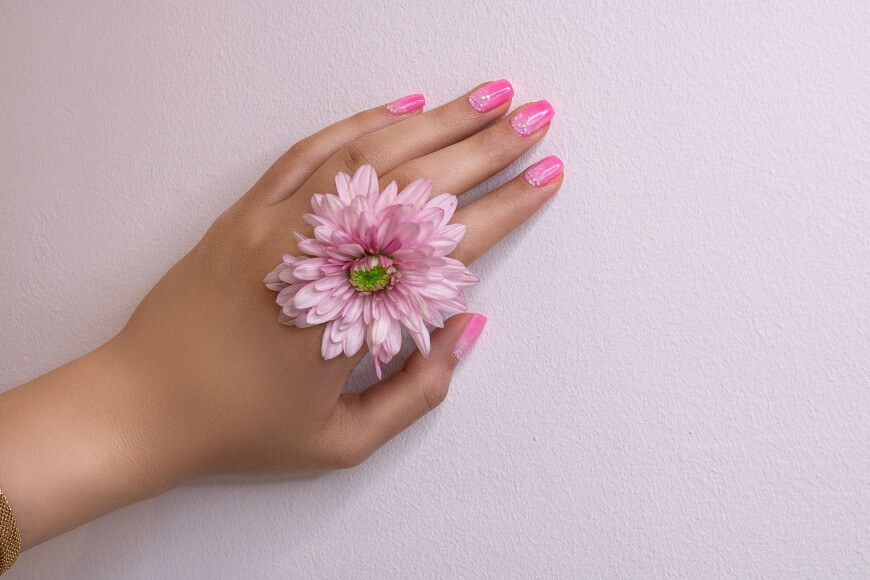 A hand with pink manicured nails rests on a light surface, adorned with a large pink flower placed between the fingers.
