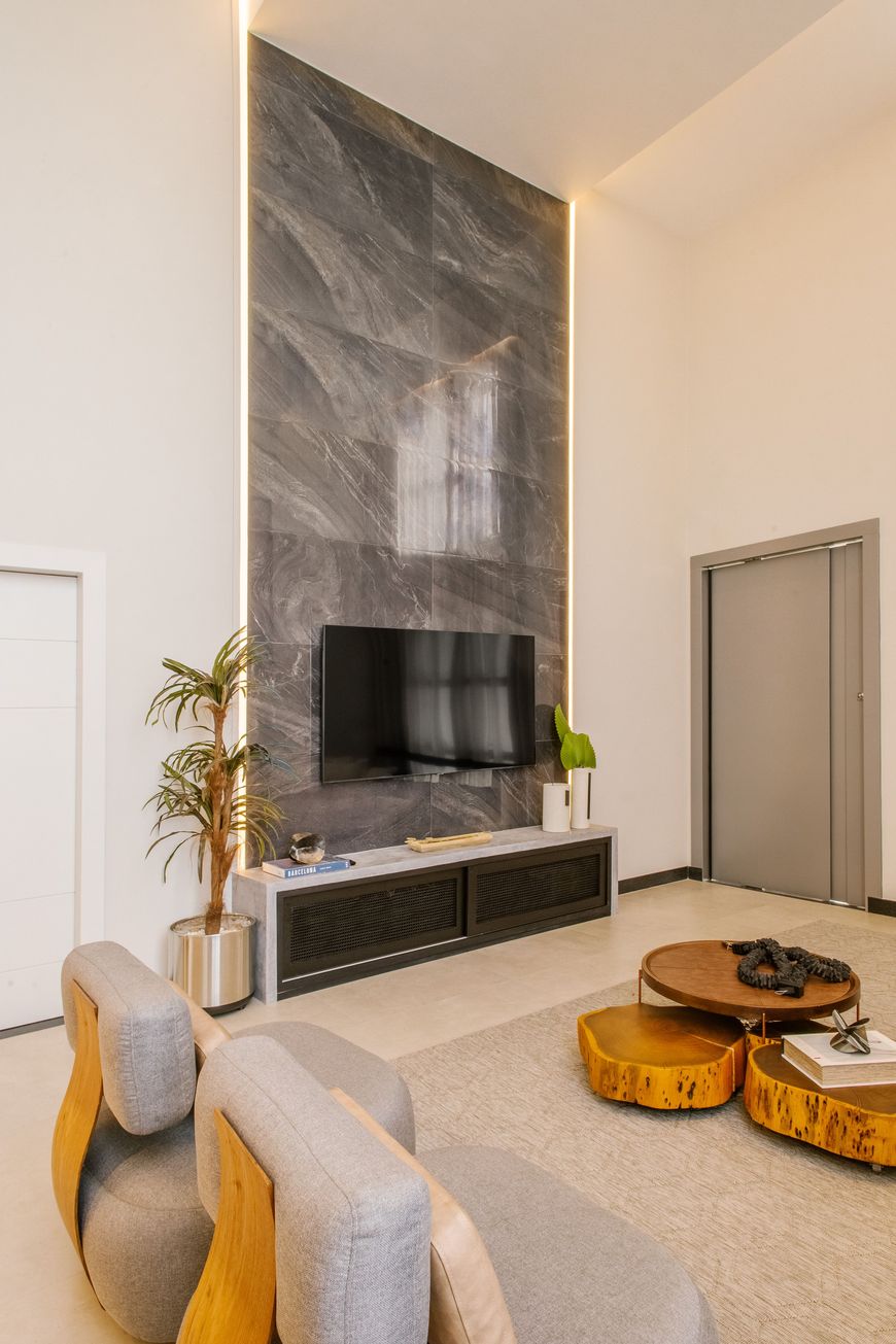 Modern living room with tall marble accent wall, mounted TV, light wood furniture, potted plants, and round wooden coffee tables on a beige rug. Natural light brightens the neutral, minimalist space.