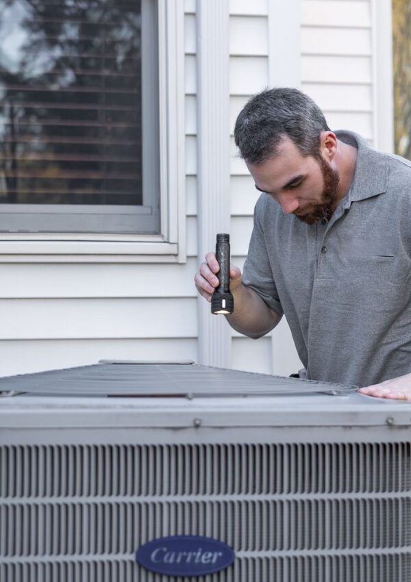 A man using a flashlight inspects an outdoor air conditioning unit next to a white house.