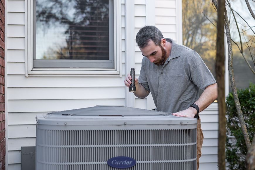 A man inspects an outdoor air conditioning unit with a flashlight near a house window, wearing a gray polo shirt and khaki pants.
