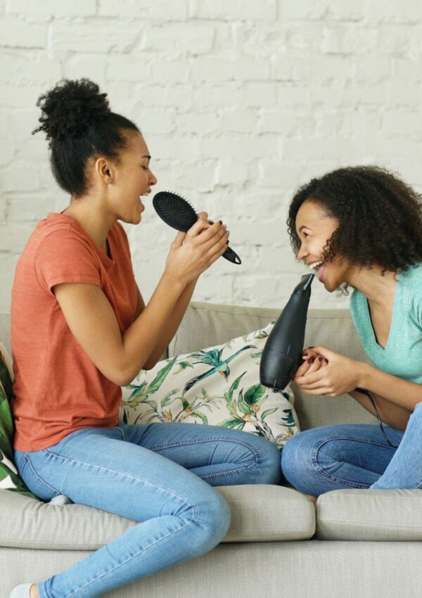 Two young women sit on a couch, smiling and pretending to sing into a hairbrush and a hairdryer, as if using them as microphones. They appear to be having fun together in a cozy living room.