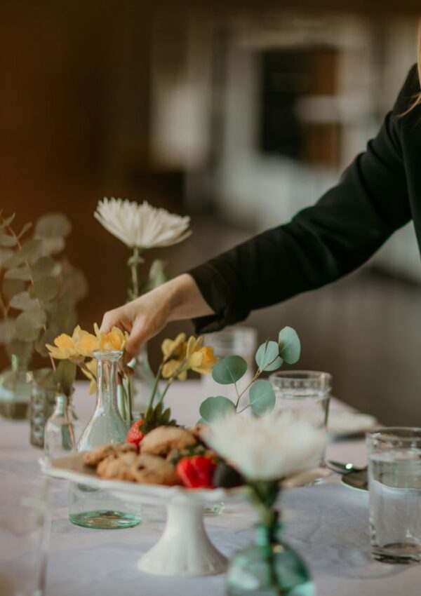 A person in a black jacket arranges yellow flowers on a table set with water glasses, cookies, berries, and small vases holding various flowers and greenery.