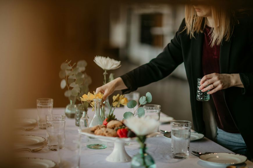 A woman arranges flowers on a table set with plates, glasses, cookies, and small vases with flowers and greenery. The scene is softly lit, creating a warm, inviting atmosphere.