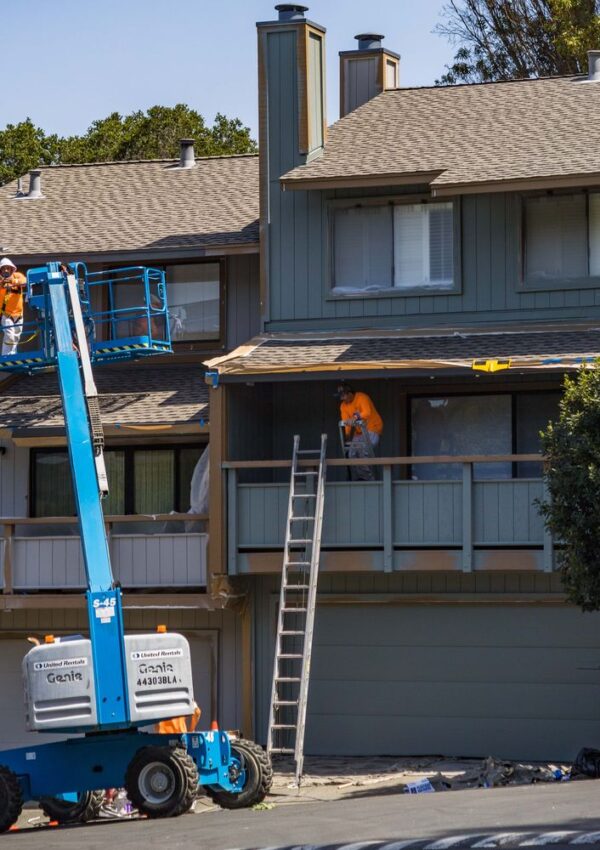 Two construction workers wearing orange shirts work on the exterior of a multi-story house. One stands on a lift, while the other is on the porch roof. A tall ladder leans against the balcony.