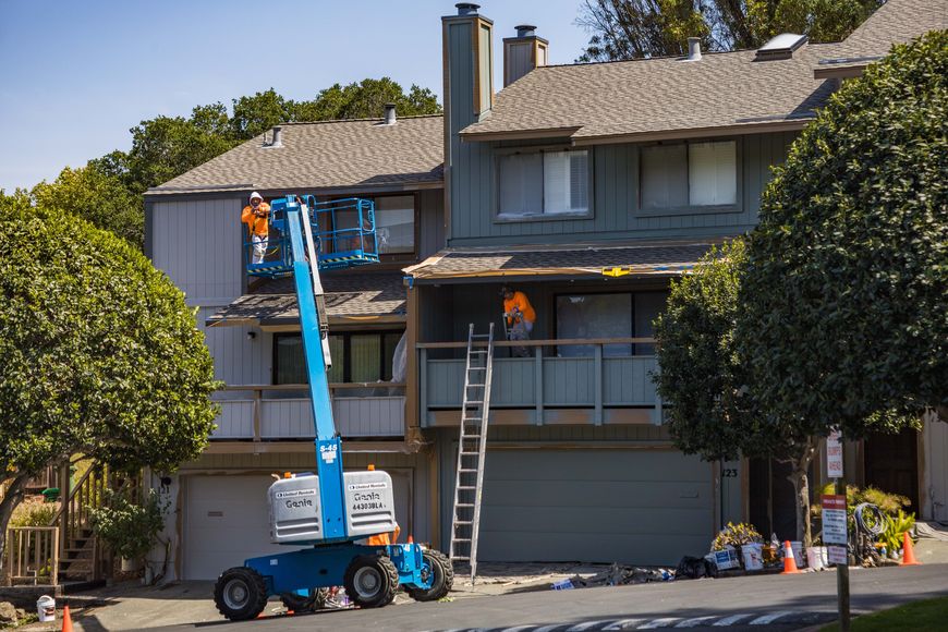 Two workers repair the roof and exterior of a two-story house using a blue lift and a ladder. Tools and supplies are scattered on the driveway below.