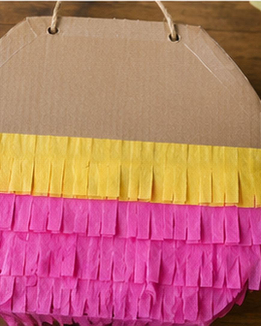 A close-up of a cardboard piñata decorated with yellow and pink fringed tissue paper, hanging by a string.