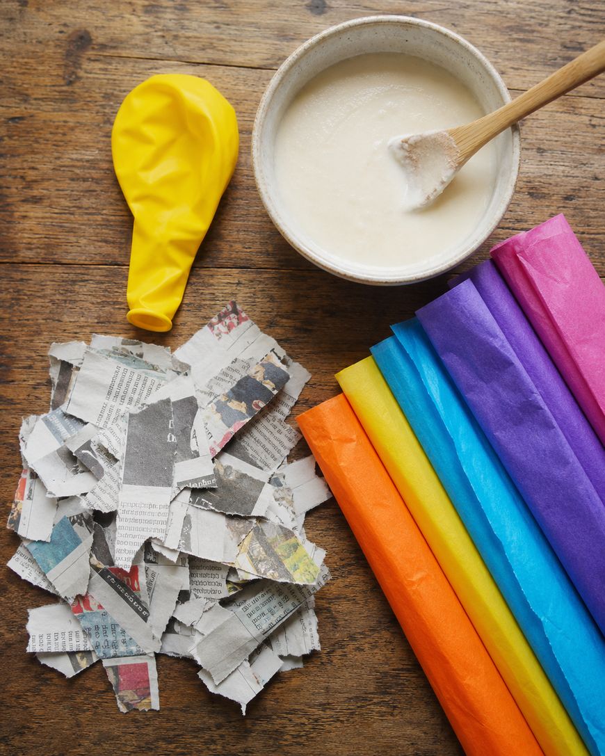 A yellow balloon, a bowl of glue with a wooden spoon, torn newspaper pieces, and colorful tissue paper strips are arranged on a wooden surface.