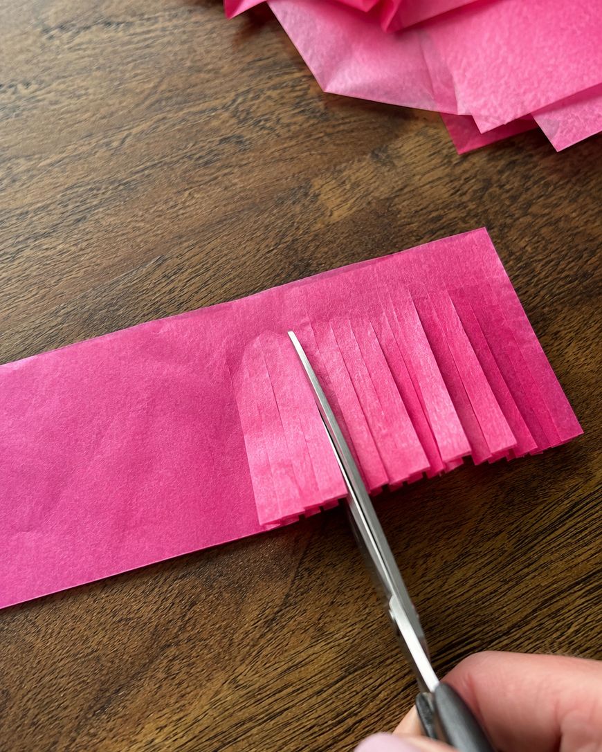 A hand uses scissors to cut fringe into a strip of pink tissue paper on a wooden surface, with more pink tissue paper sheets in the background.