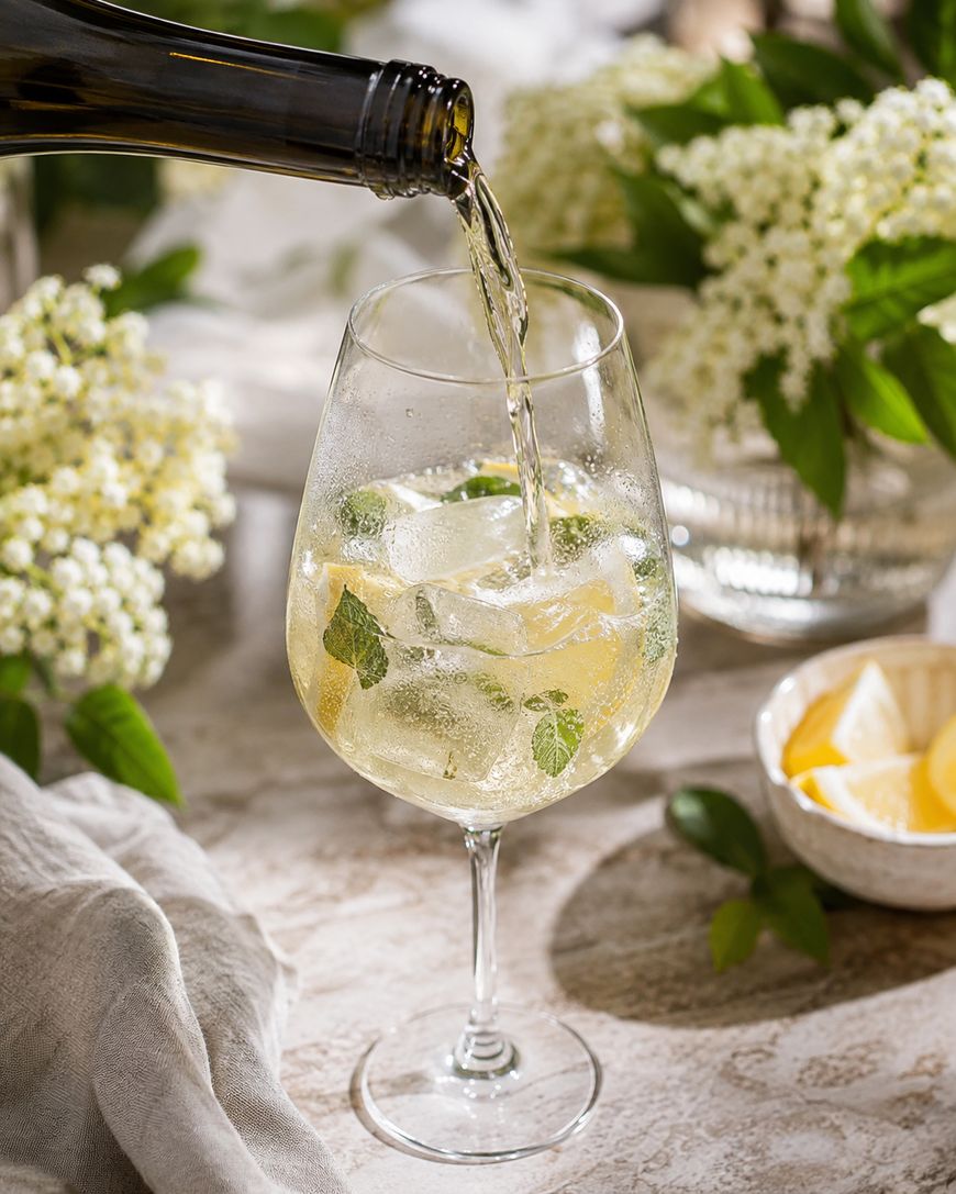 A glass of white wine with ice cubes and mint leaves is being poured, surrounded by elderflower blossoms, a bowl of lemon wedges, and a light tablecloth on a bright, elegant table.