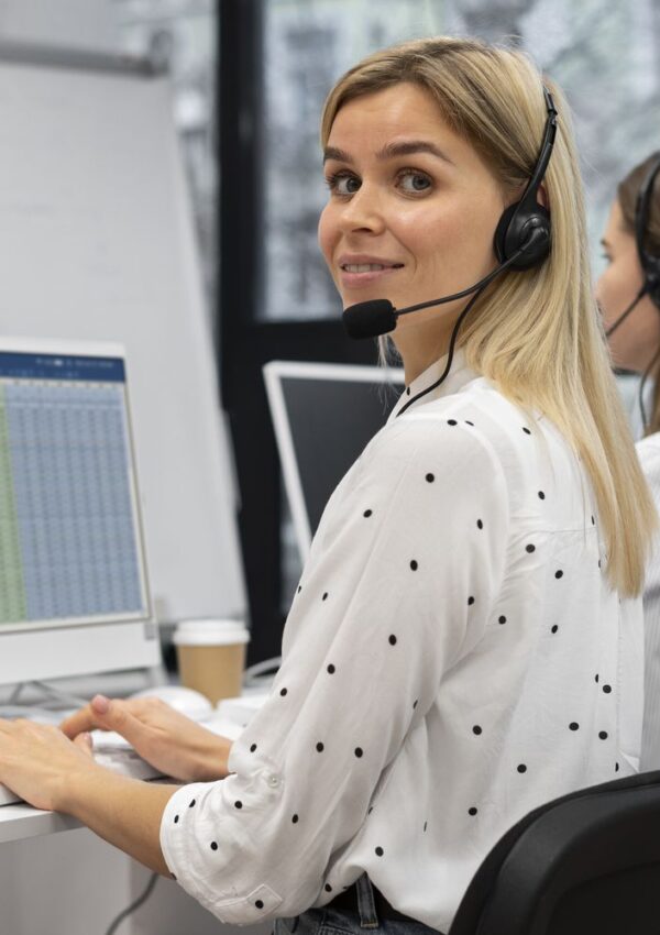 A woman with a headset sits at a desk, smiling at the camera, working on a computer with a spreadsheet on the screen. Another woman with a headset is visible in the background.