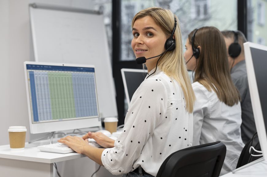 A woman wearing a headset sits at a desk in an office, smiling at the camera, with a computer monitor displaying a spreadsheet. Two colleagues are working beside her. Coffee cups are on the desks.