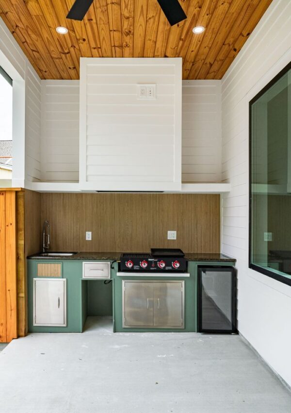 Modern outdoor kitchen with green cabinets, a grill, small sink, mini fridge, and wooden ceiling. Large window on the right and wood panel wall on the left. Clean, minimalist design with neutral colors.