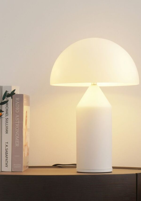 A modern white table lamp with a dome-shaped shade sits on a dark wooden surface next to three books and a white vase containing orange flowers. The lamp is turned on, casting a warm light.