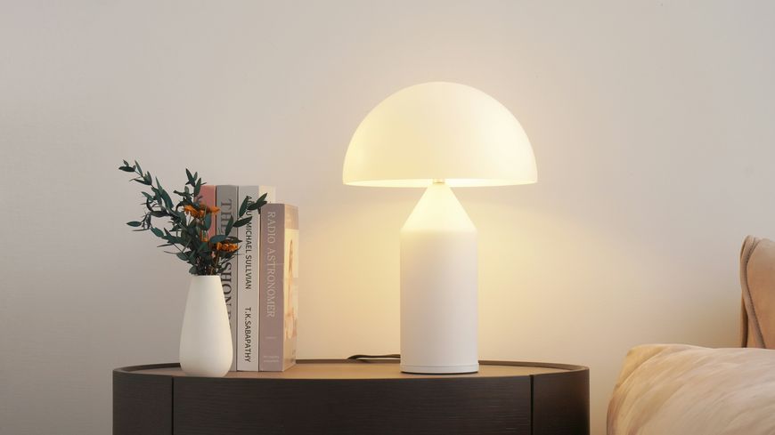 A round side table with a white vase of flowers, three books, and a modern white table lamp with a dome-shaped shade, softly illuminating the space.