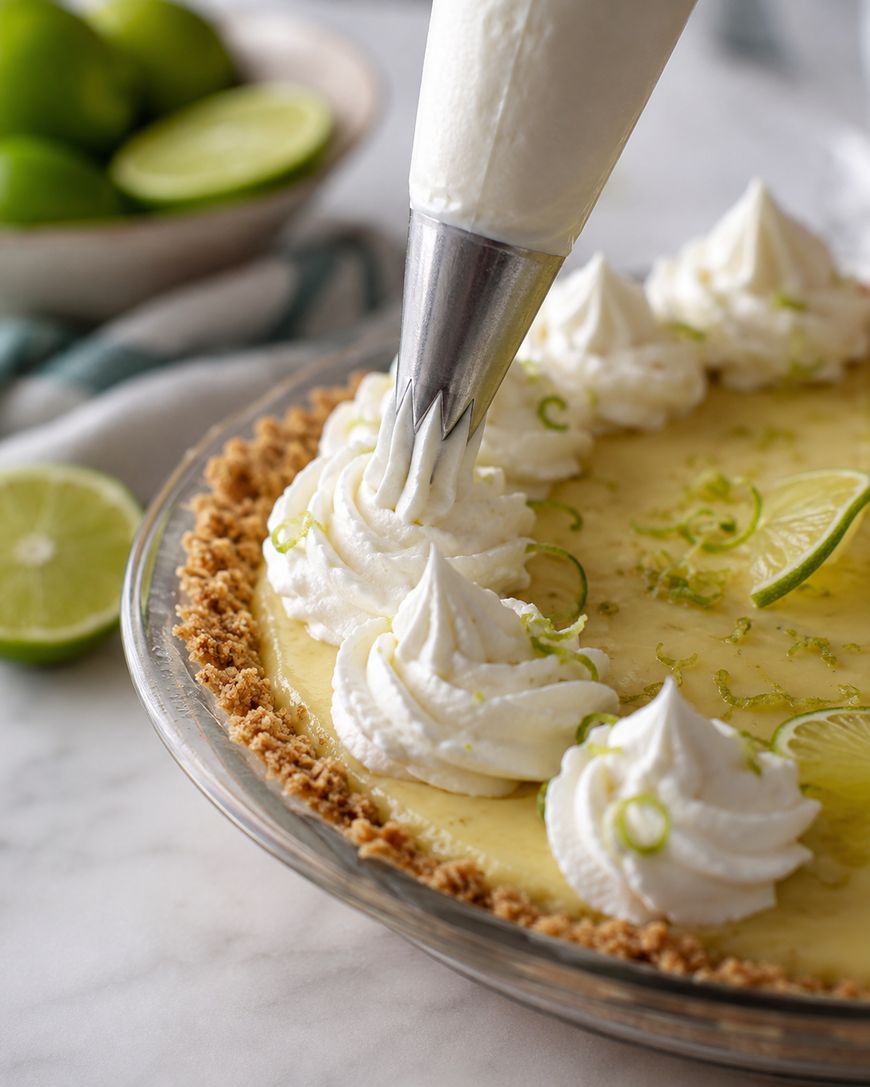 A close-up of a key lime pie being decorated with whipped cream from a piping bag. Slices of lime and lime zest are on top, with whole limes and a blue towel in the background.