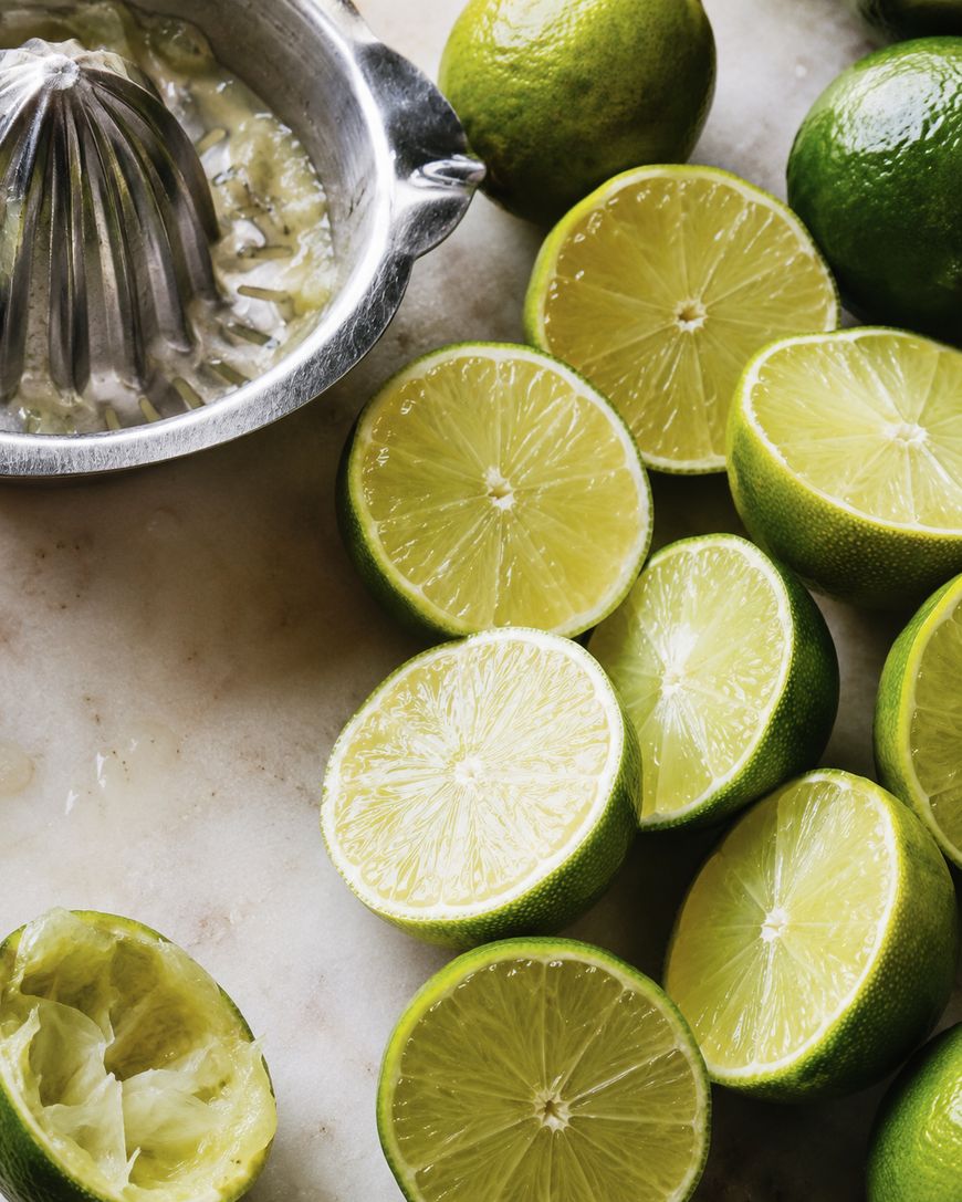 Halved limes are arranged on a surface next to a metal citrus juicer with squeezed lime halves, showing fresh, juicy interiors.
