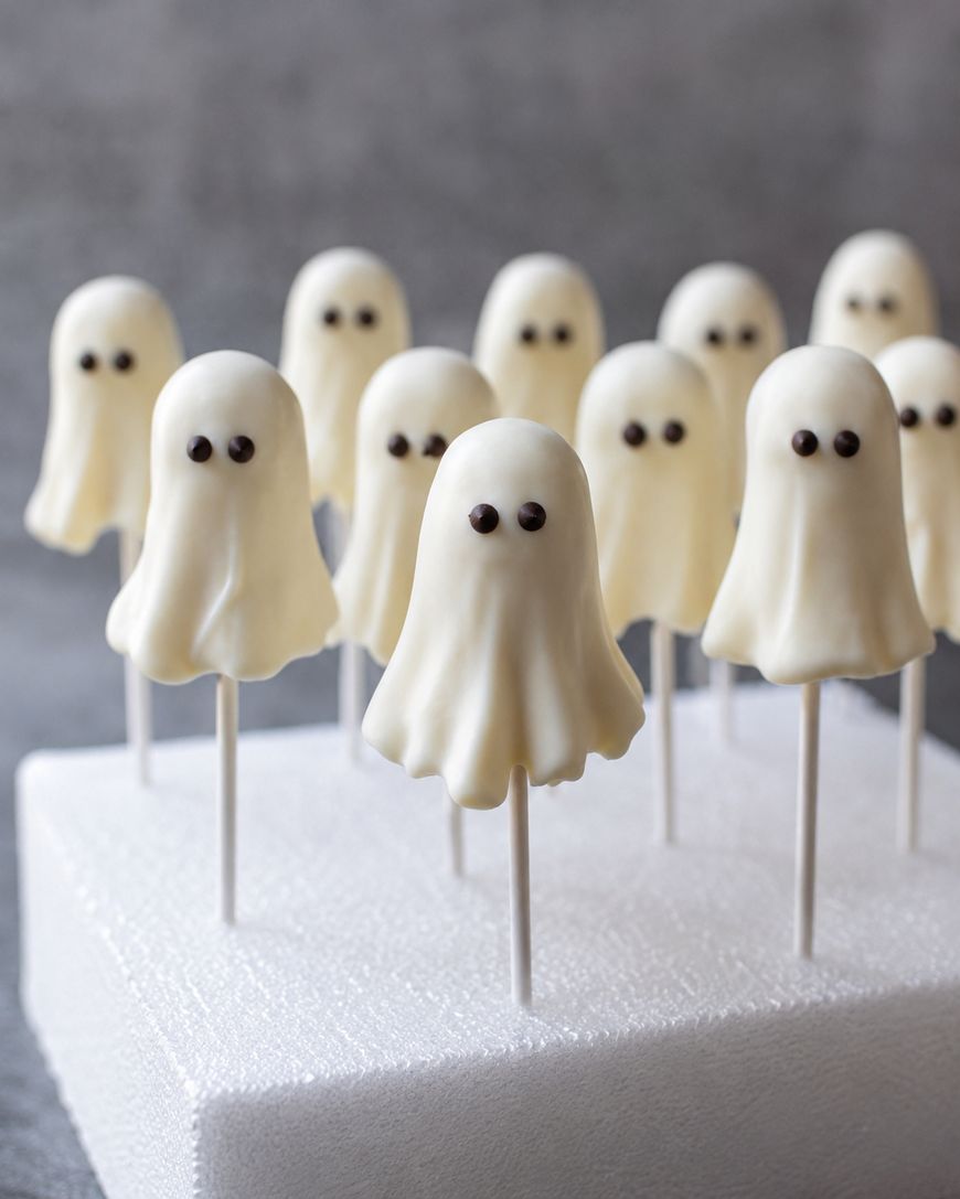 Twelve cake pops decorated as cute white ghosts with black eyes, standing upright on sticks in a white foam block, with a soft gray background.