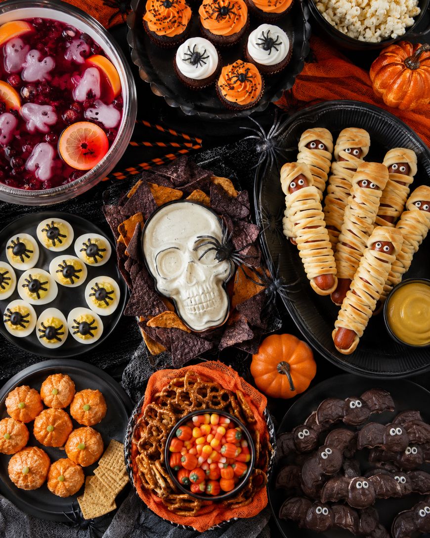 A Halloween-themed table with skull dip, mummy hot dogs, spider cupcakes, eyeball deviled eggs, popcorn, cookies, punch, mini pumpkins, and candy corn, all arranged festively with orange and black decorations.