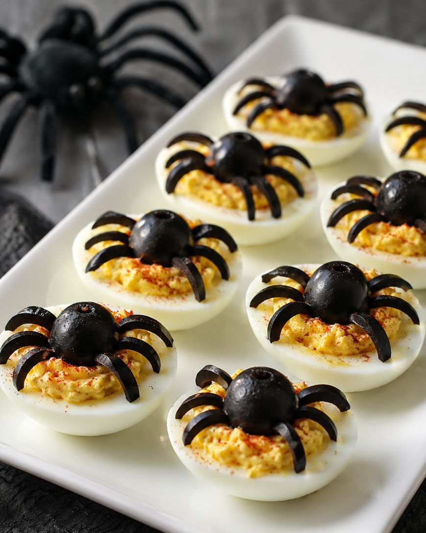 Deviled eggs topped with black olives cut to resemble spiders, arranged on a white rectangular plate. A black plastic spider decoration is in the background.