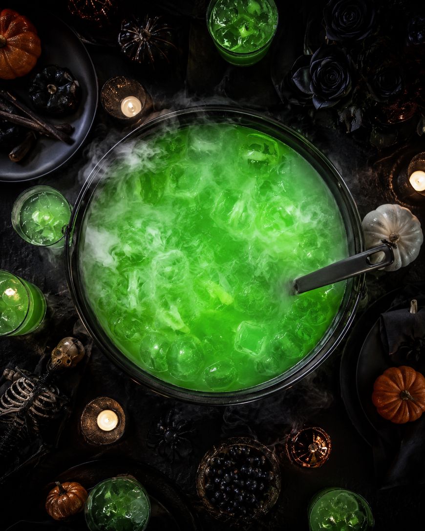 A large cauldron of bright green, smoking punch surrounded by candles, black roses, pumpkins, skeleton decor, and glasses of the same drink, creating a spooky Halloween party setting.