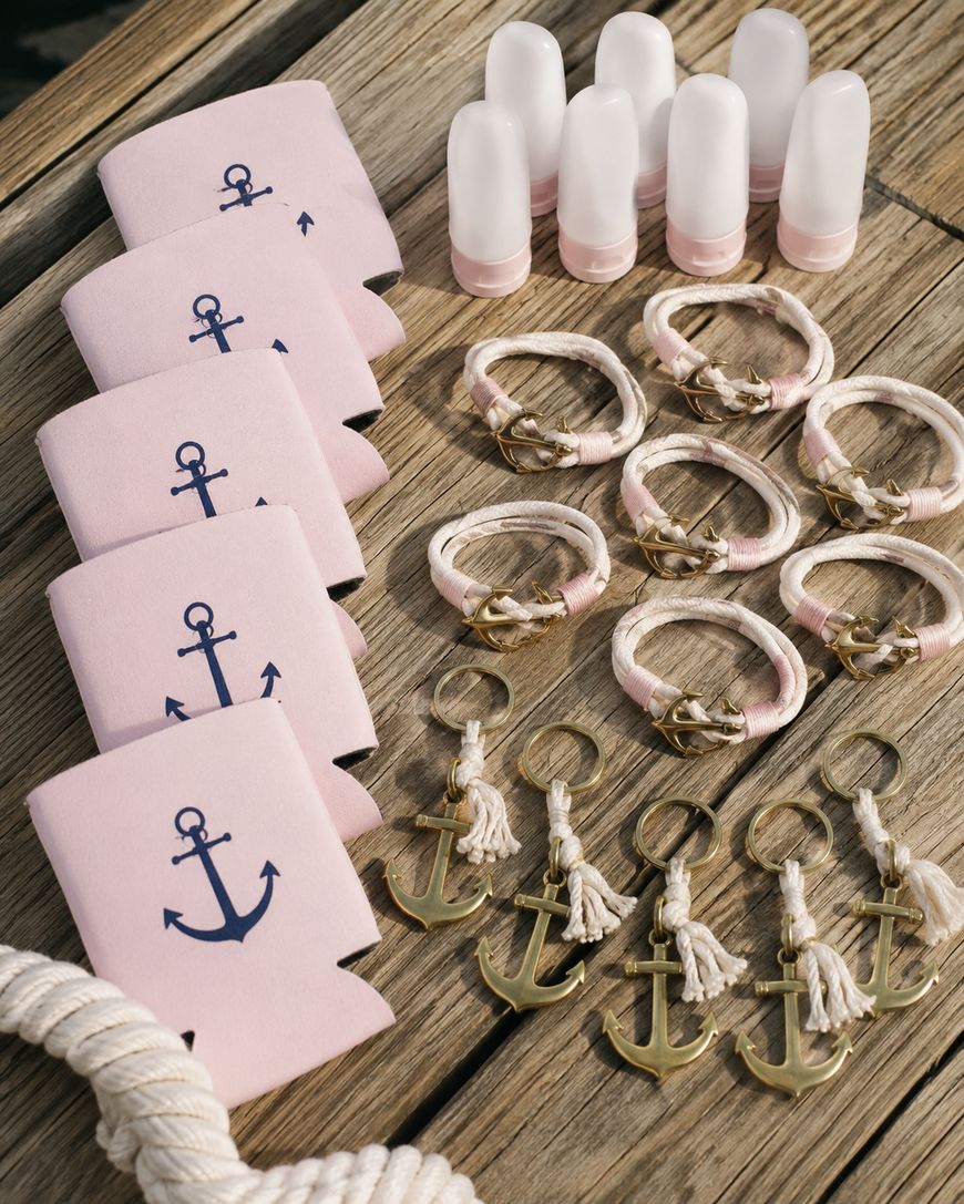Pink anchor koozies, travel bottles, braided rope bracelets with anchors, and anchor keychains arranged neatly on a wooden surface, suggesting a nautical or beach-themed event or party.