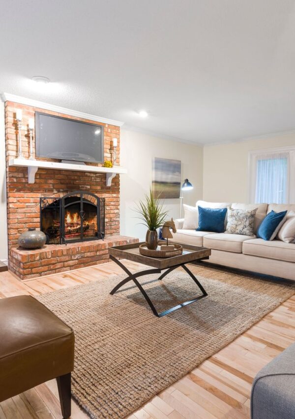 A cozy living room with a brick fireplace, TV above mantel, beige sofa with blue pillows, modern coffee table, area rug, hardwood floor, and soft lighting.