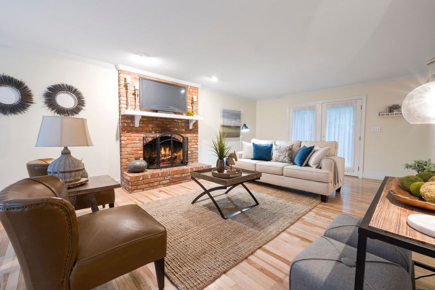 Bright living room with a brick fireplace, TV, beige sofa, brown leather chairs, coffee table, rug, and decorative accents on hardwood floors. Light streams in through French doors in the background, highlighting thoughtfully chosen living room decor that adds home personality to the inviting space.