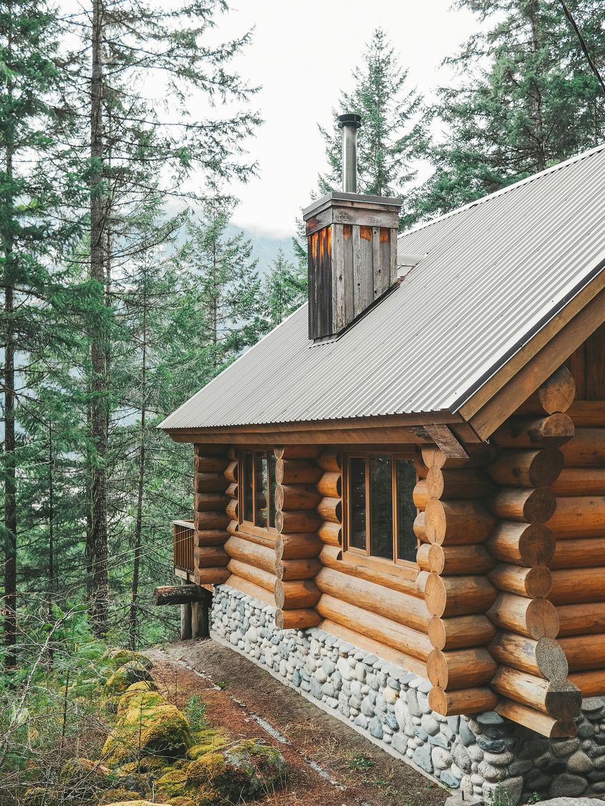 A cozy log cabin with a stone foundation and metal roof sits among tall evergreen trees in a forest. Designed to maximise space, this charming retreat showcases the flexible interiors often found in Log Cabins Ireland. Smoke rises gently from the chimney, promising warmth and comfort within.
